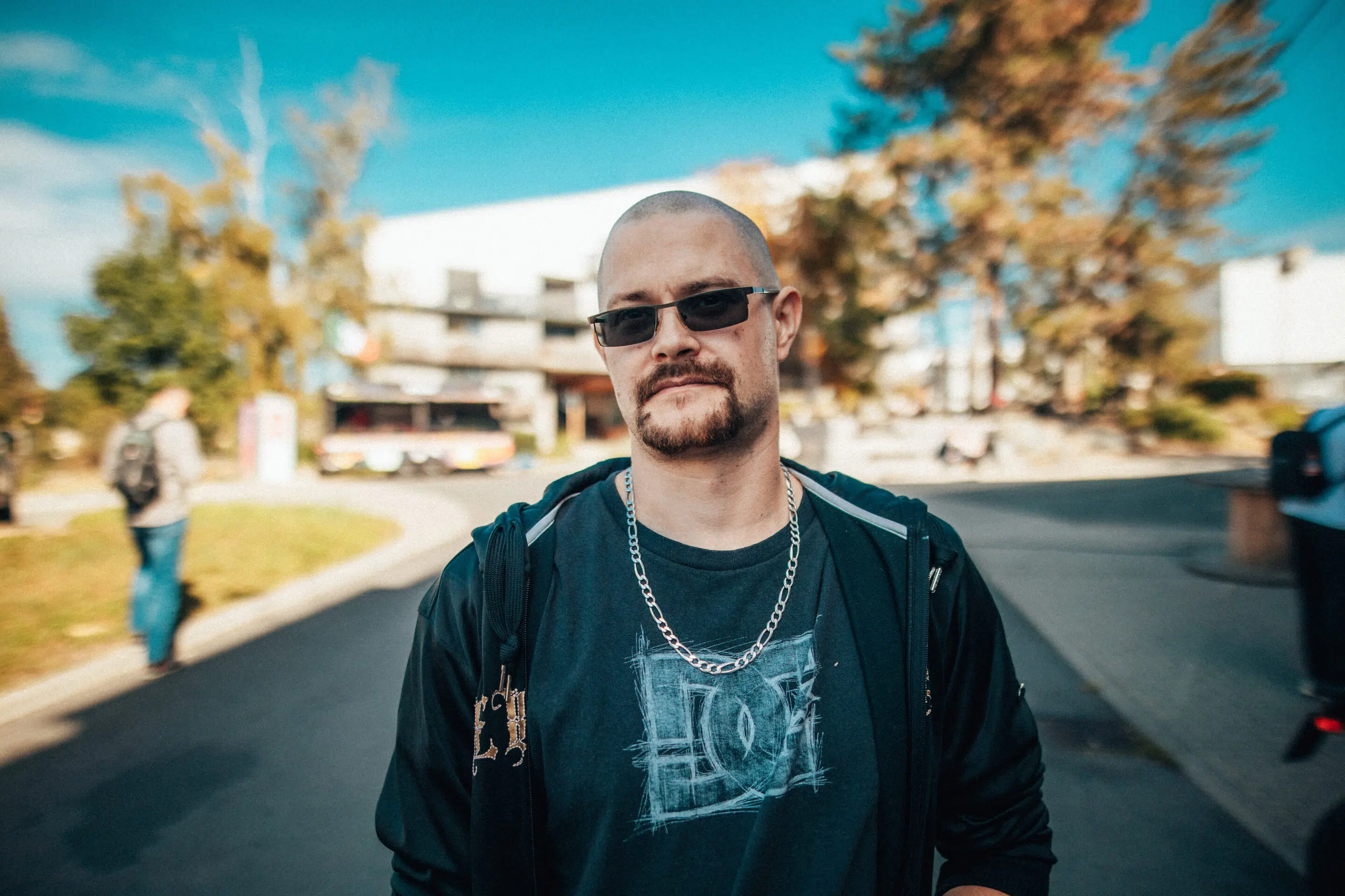 Lukáš Kenji Vrábel - www.kenji.cz Muž s krátkými vlasy a slunečními brýlemi, oblečený v černé bundě, tričku s nápisem a řetízkovém náhrdelníku, stojí venku za slunečného dne se stromy a budovami v pozadí.