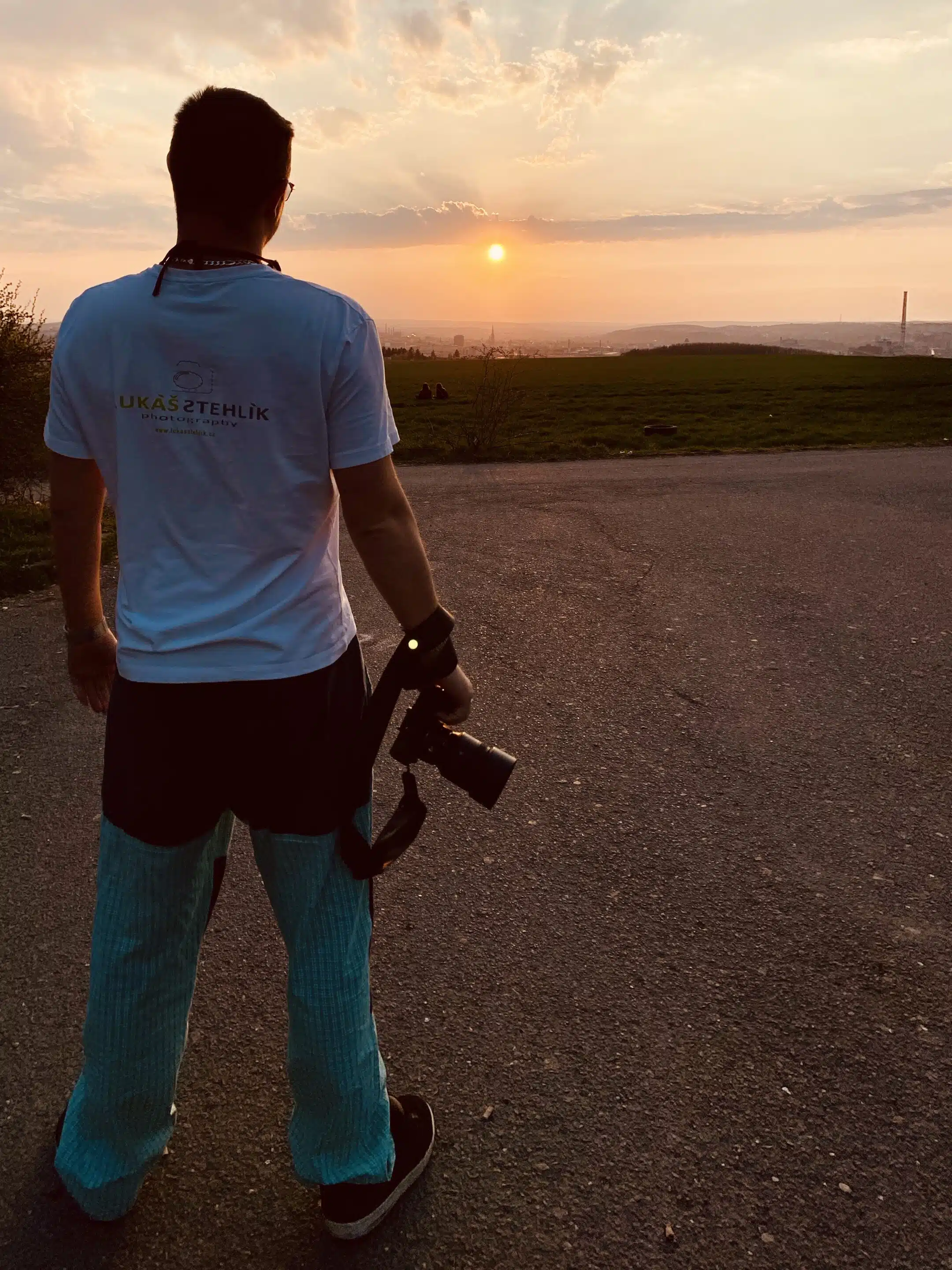 lukasstehlik-00006 Člověk stojí na dlážděné cestě při západu slunce, drží fotoaparát a dívá se k horizontu, se zelenou trávou a vzdálenou siluetou města.