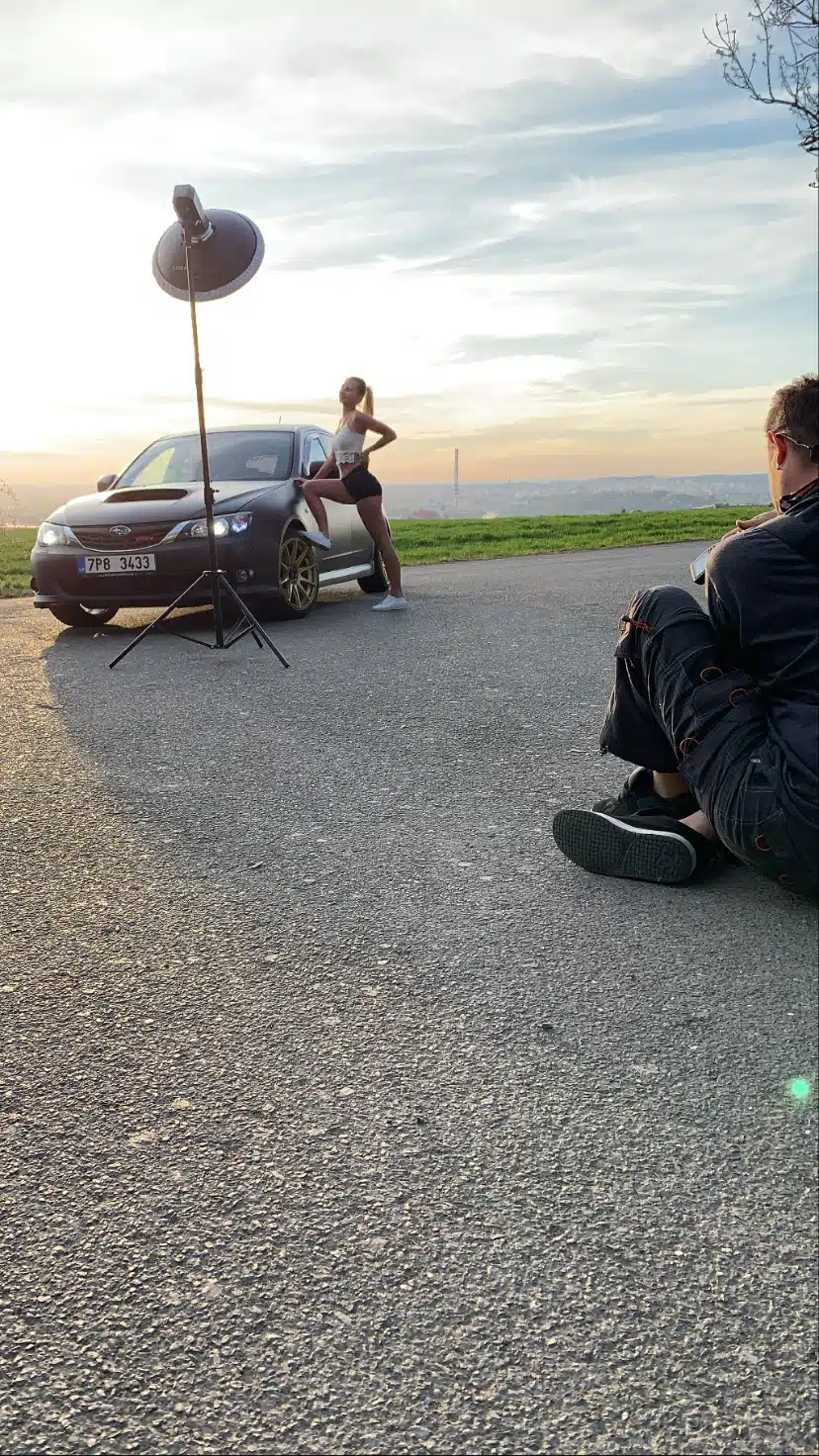 lukasstehlik-00007 Žena pózuje vedle zaparkovaného auta během focení, fotograf sedí na zemi a poblíž je při západu slunce rozsvíceno studiové světlo.