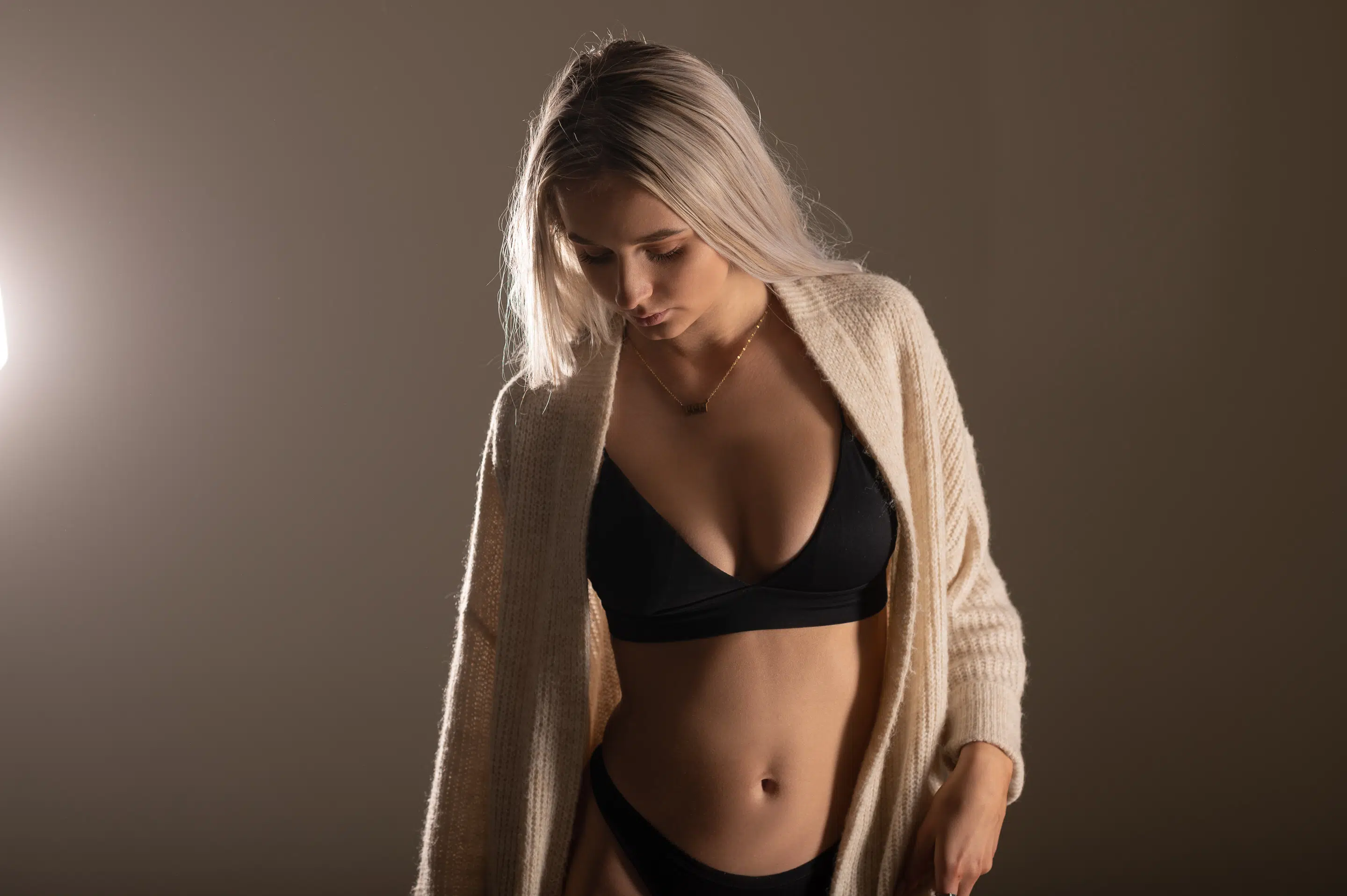 Žena s dlouhými blond vlasy má na sobě krémový kardigan přes černou podprsenku a spodní prádlo a stojí na neutrálním pozadí s měkkým osvětlením.